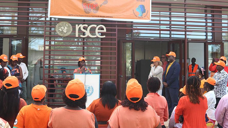 The backs of people all wearing orange facing a stage where a young woman in an orange baseball cap is addressing the crowd.