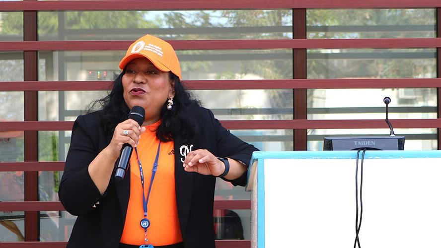 A woman wearing an orange baseball cap and orange shirt speaking into a microphone to address an audience.