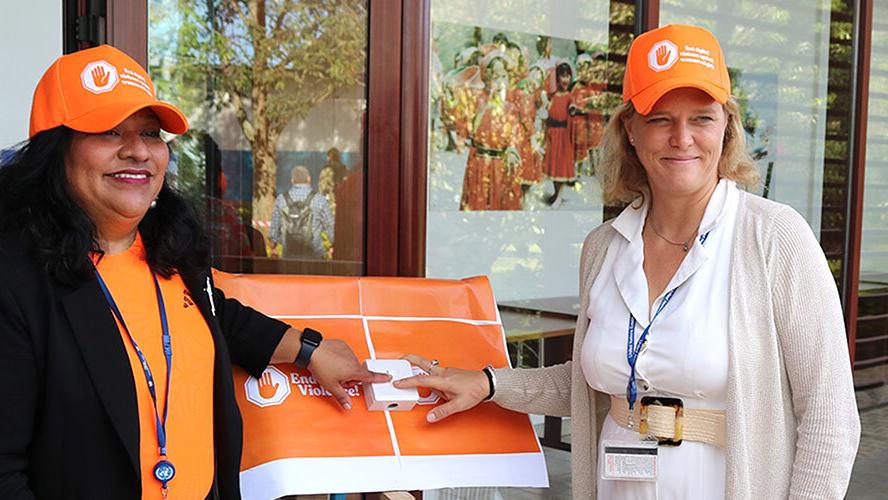 Two women both wearing orange coloured clothing and smiling each putting a finger on a button between them