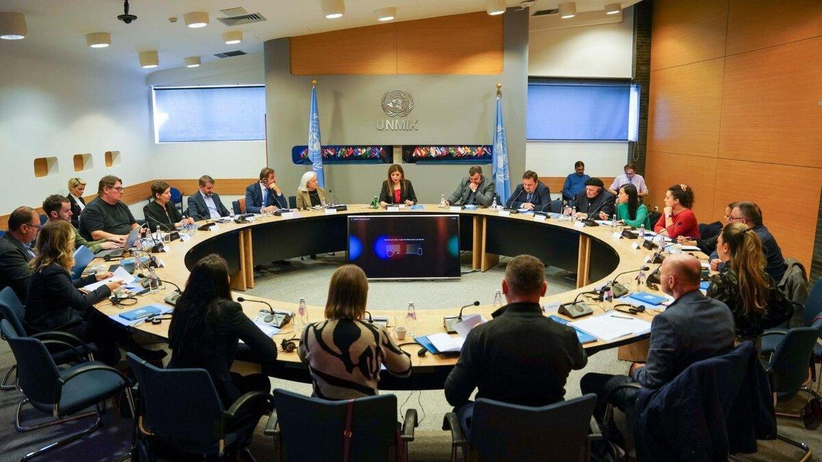 Large roundtable meeting with multiple participants seated around a curved conference table. UNMIK logo and two UN flags are visible in the background, with documents, microphones, and water bottles on the table.