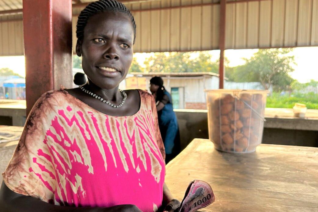 Person standing at a market stall holding currency notes, with a container of fried snacks on the counter and open-air structures in the background.