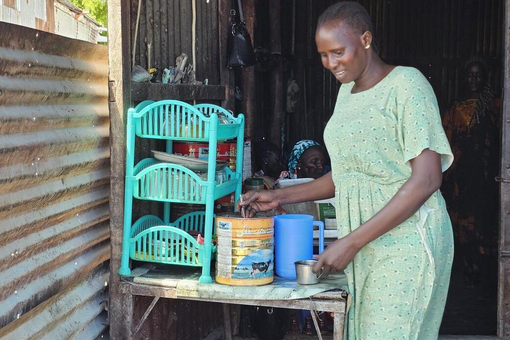 A person in a light green dress stands at a small table inside a corrugated metal structure, preparing items near plastic shelves and containers.