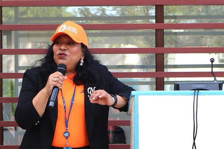 A woman wearing an orange baseball cap and orange shirt speaking into a microphone to address an audience.