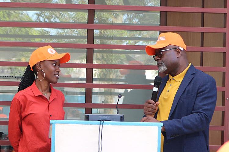 A young woman in an orange baseball cap smiling and speaking to a man also wearing an orange baseball cap. 